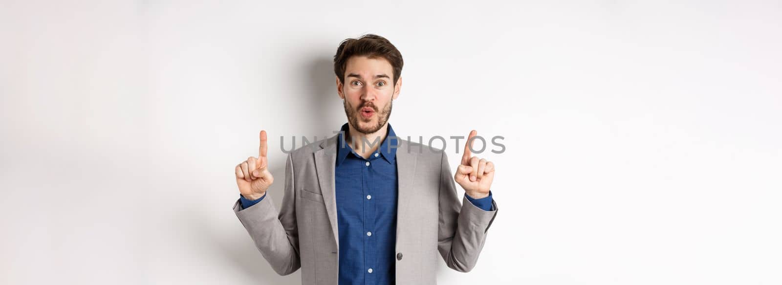 Excited businessman in suit say wow and smiling amused, pointing fingers up at good deal, standing against white background.