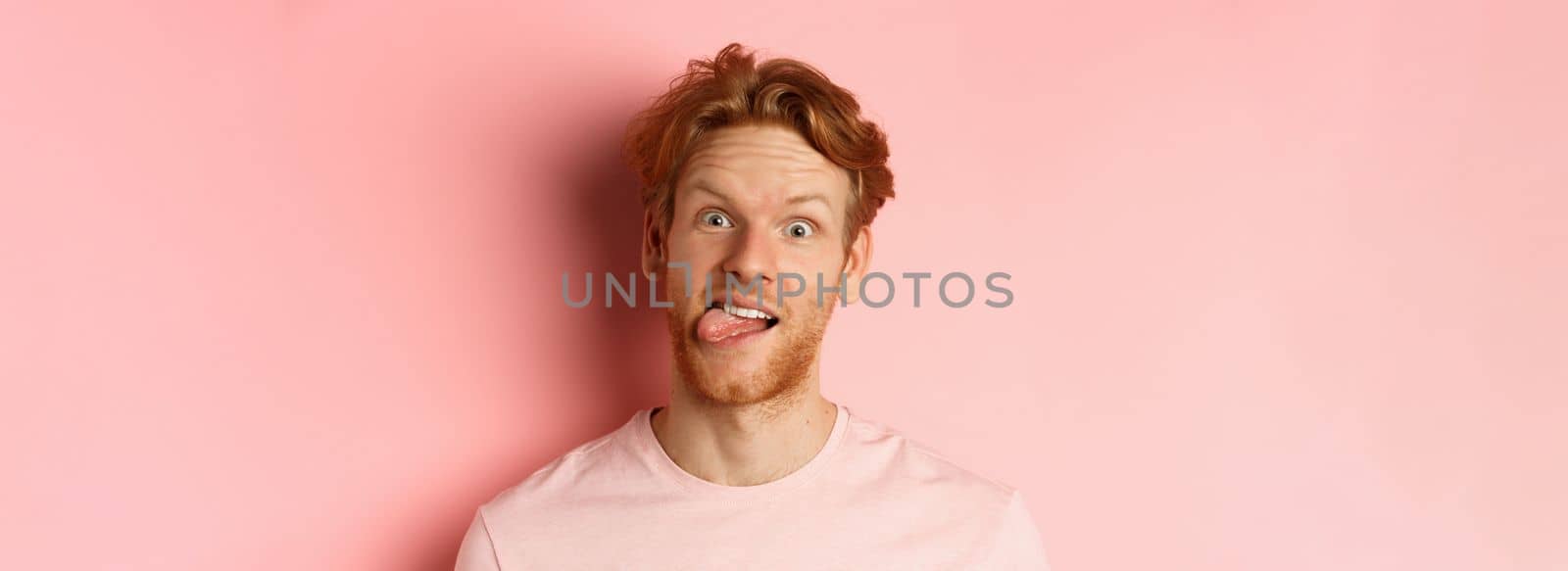 Headshot of funny redhead guy showing tongue, making silly faces at camera, standing joyful against pink background by Benzoix