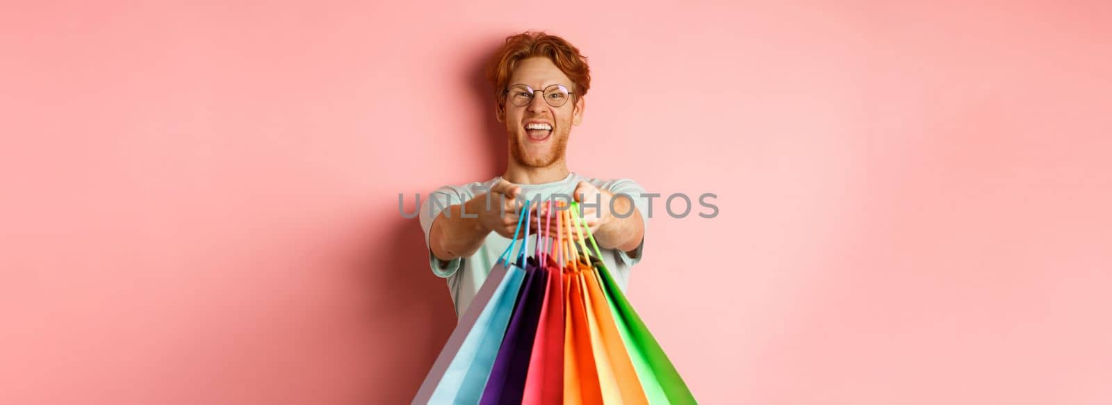 Happy redhead man stretch out hands with shopping bags, give you gifts, standing over pink background by Benzoix