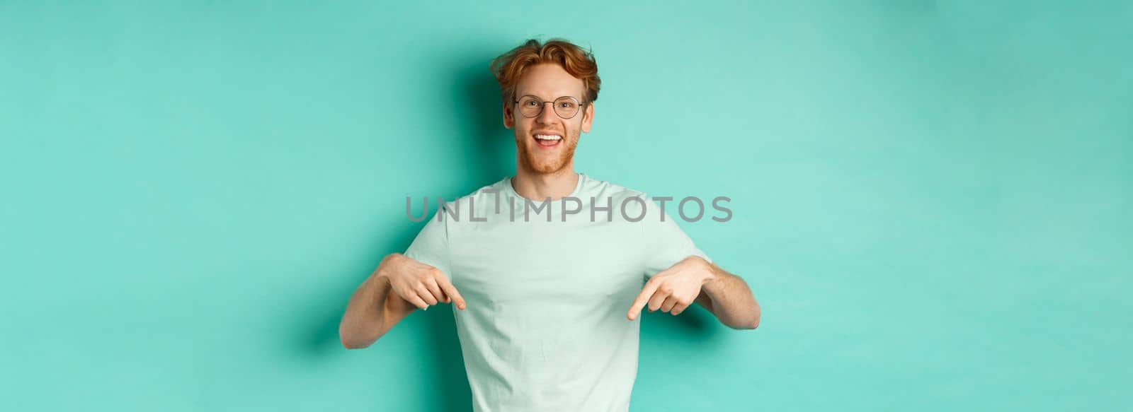 Happy young man with red hair, wearing glasses and t-shirt, showing advertisement, smiling and pointing fingers down, standing over turquoise background.