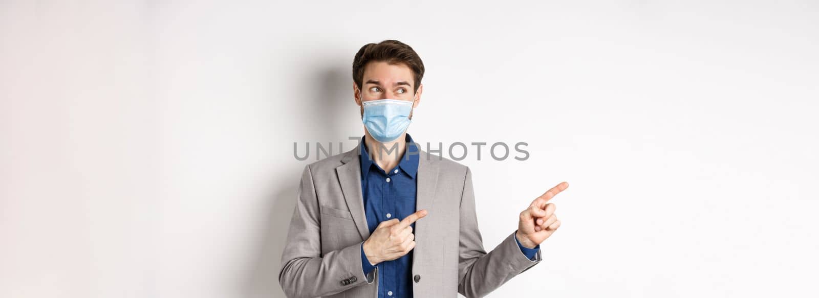 Covid-19, pandemic and business concept. Excited young man in medical mask and suit looking left, pointing at logo with pleased face, white background.