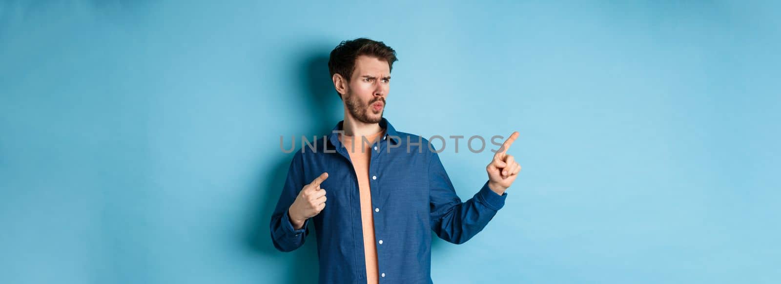 Confused and annoyed guy pointing and looking left at empty space with angry frowning face, standing on blue background.