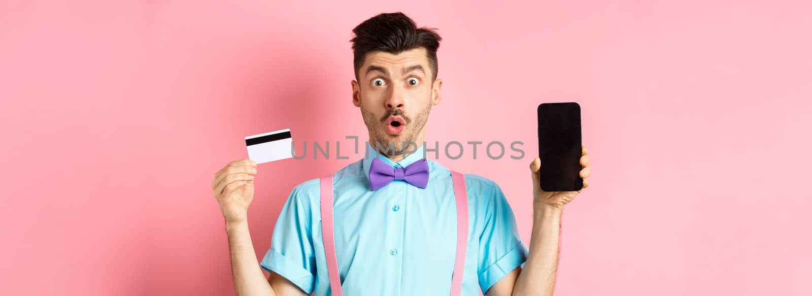 E-commerce and shopping concept. Surprised man showing blank smartphone screen with plastic credit card, standing amazed on pink background.