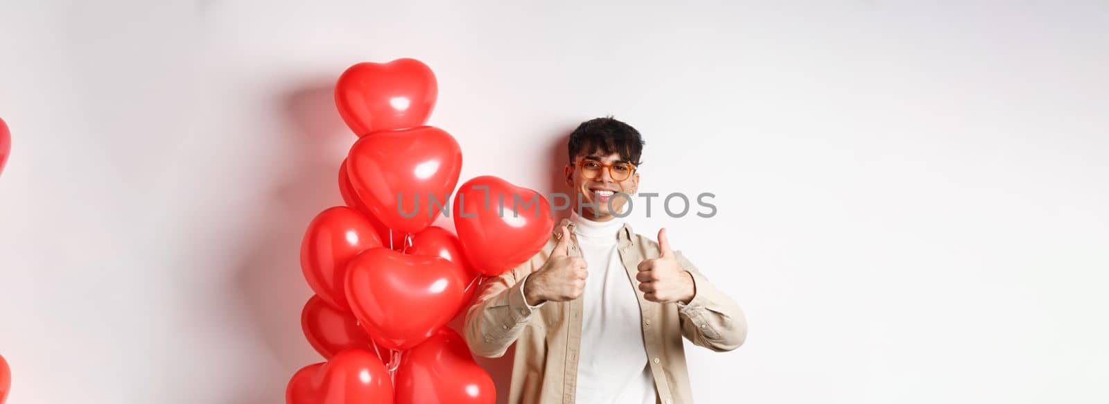 Valentines day and romance concept. Stylish modern man in sunglasses showing thumbs up while standing near heart balloons, saying yes, enjoy date with lover by Benzoix