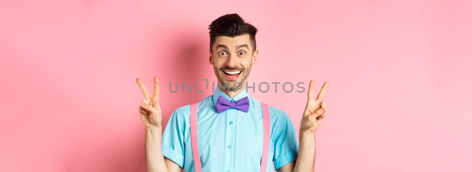 Cheerful young guy with moustache and bow-tie showing peace or victory gestures, smiling happy at camera, standing on pink background.