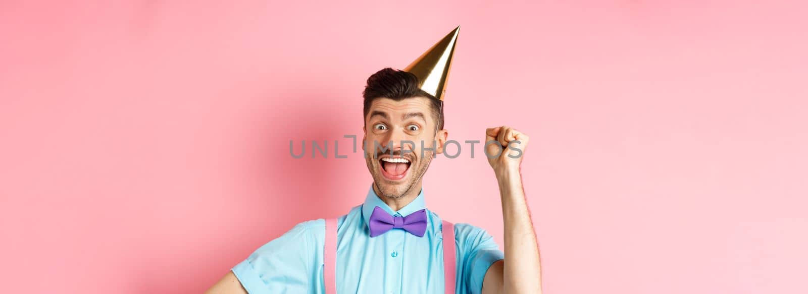 Holidays and celebration concept. Happy birthday boy with moustache and bow-tie celebrating event in party cone hat and festive clothes, raising hand up and shout yes.