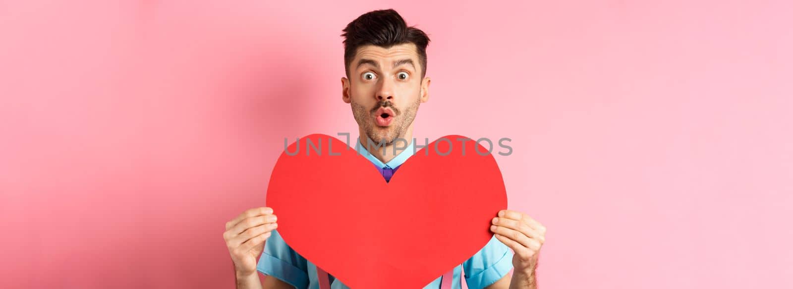 Valentines day concept. Amused young man with bow-tie and moustache, showing heart cutout and looking for love, standing on pink romantic background.