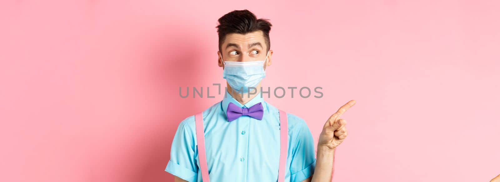 Covid-19, pandemic and health concept. Funny man in bow-tie and medical mask pointing finger left, looking aside curious, showing logo, standing on pink background.