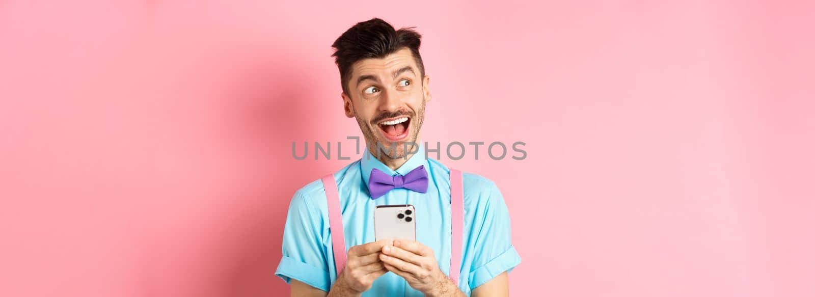 Technology concept. Excited man looking aside with dreamy face, winning online prize on smartphone, standing cheerful over pink background.