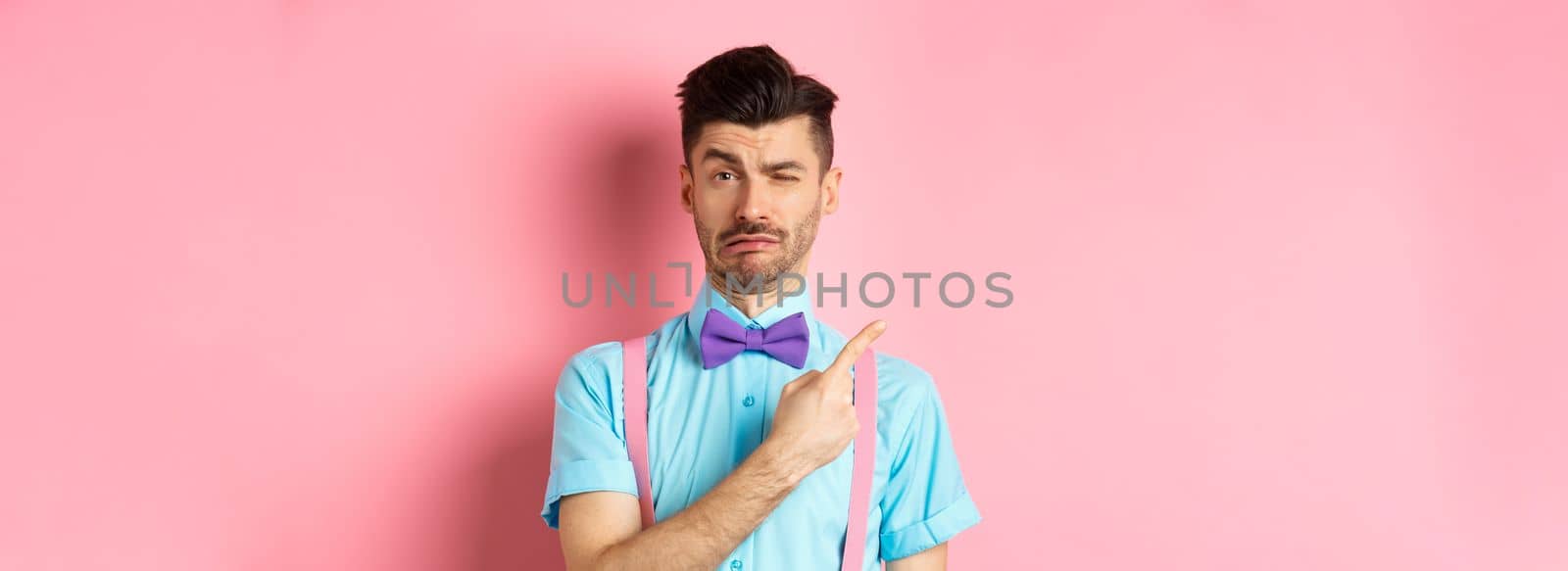 Childish young man acting sad and fake crying while pointing finger upper left corner, complaining on something, standing over pink background by Benzoix