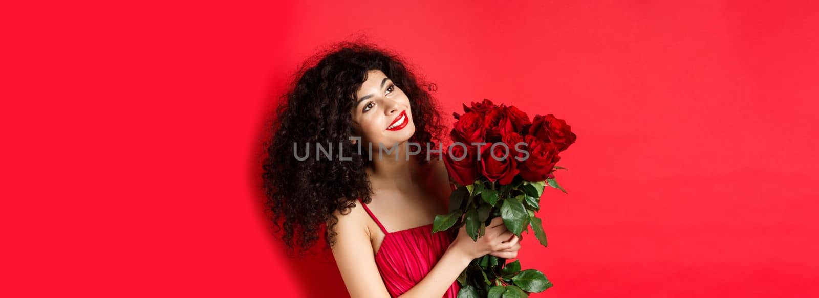 Happy beautiful woman in dress, holding flowers and smiling romantic, looking aside at logo, standing against red background by Benzoix