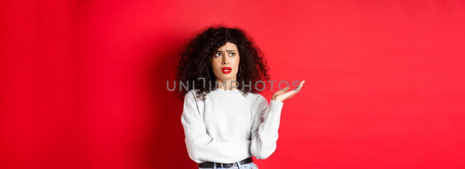 Annoyed and puzzled young woman with curly hair, raising hands up and looking aside, cant understand something strange, standing on red background.