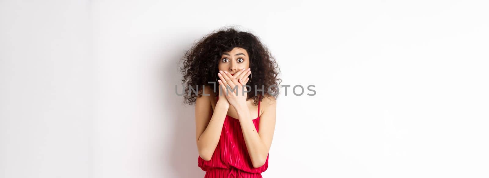 Shocked woman covering mouth with hands and staring with disbelief at camera, witness something shocking, standing in red dress on white background by Benzoix