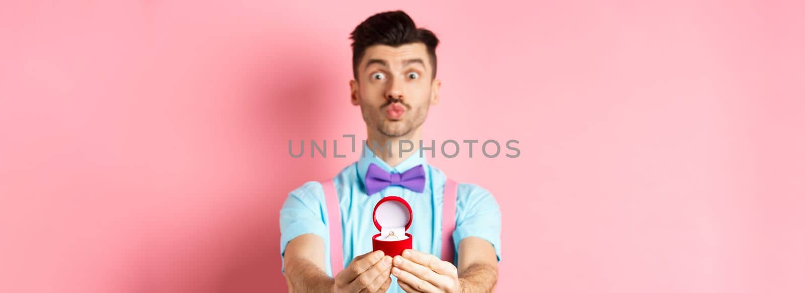 Valentines day. Funny young man pucker lips for kiss and showing engagement ring, making proposal, say marry me to lover, standing over pink background.