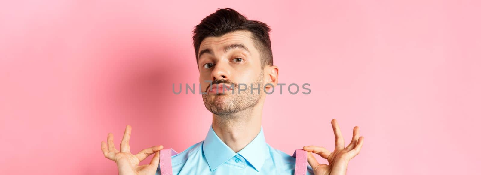Close up of classy handsome guy adjusting suspenders, pucker lips and raise chin up, looking confident, standing in trendy outfit on pink background.