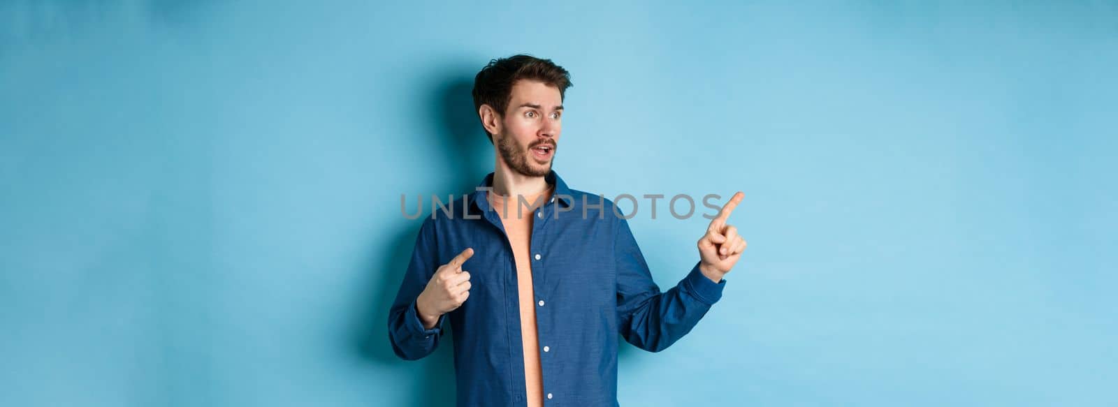 Amazed caucasian guy pointing and looking left, checking out advertisement, standing on blue background.