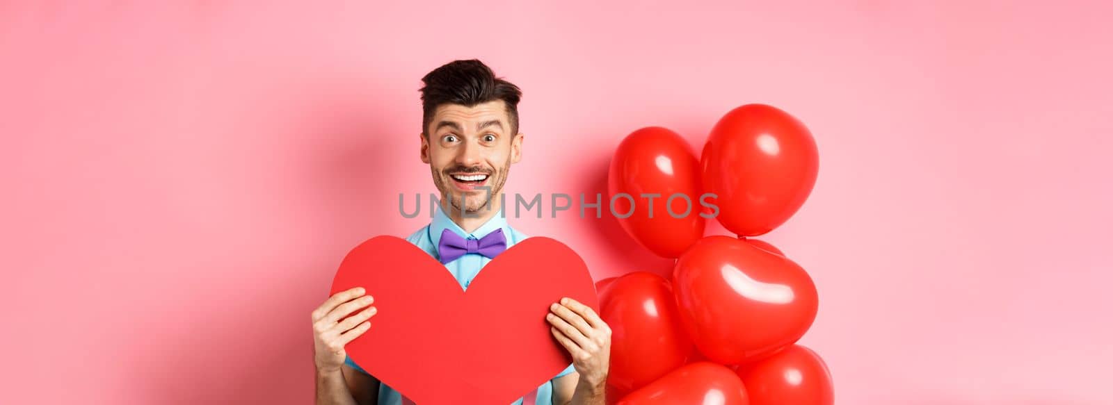 Valentines day concept. Cute young man in bow-tie showing big red heart postcard and say love you, smiling happy at camera, standing on romantic pink background by Benzoix