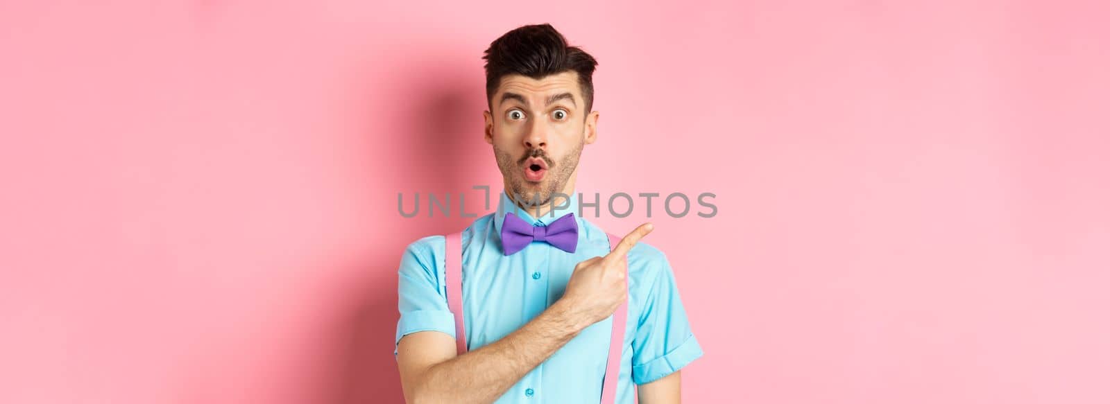 Check it out. Amazed caucasian man with moustache, wearing bow-tie, saying wow and pointing left, showing advertisement, standing on pink background.