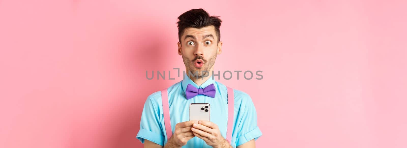 Online shopping. Intrigued guy in bow-tie, checking out internet promo offer on mobile phone, say wow at camera, holding smartphone, standing on pink background.