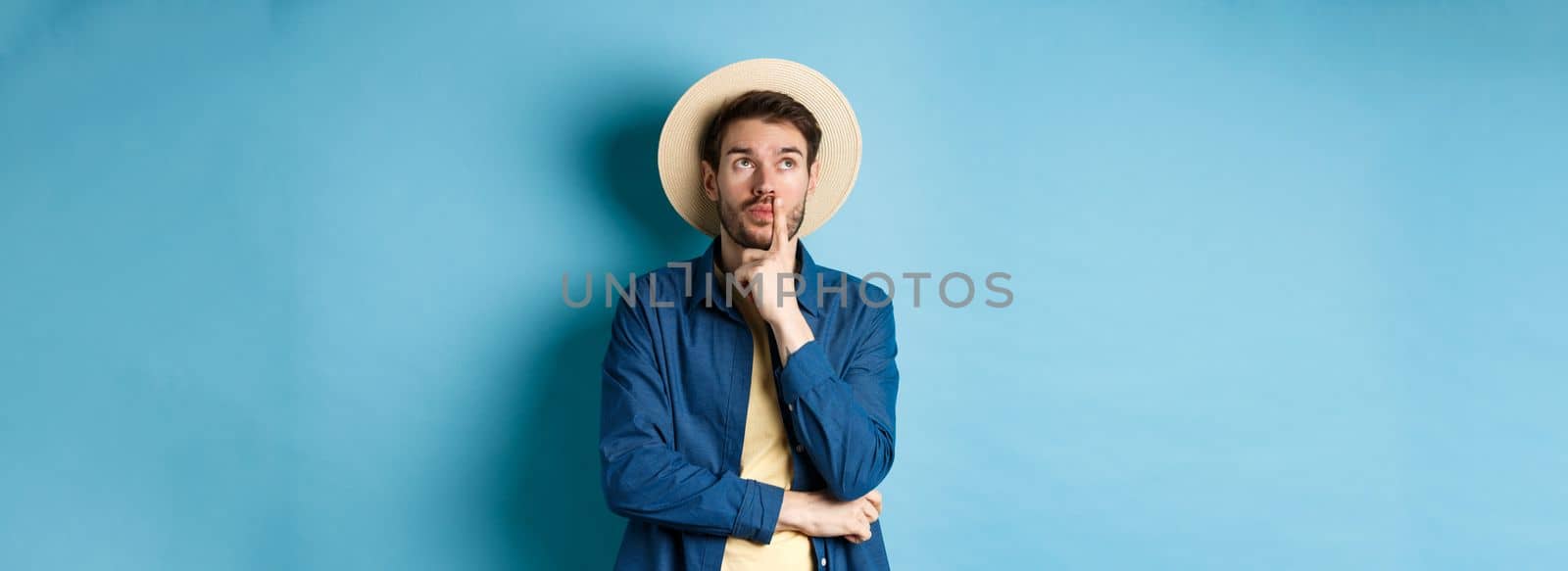 Thoughtful male tourist thinking of summer holiday vacation, looking aside with pensive face, touching lip and pondering next travel, standing on blue background.