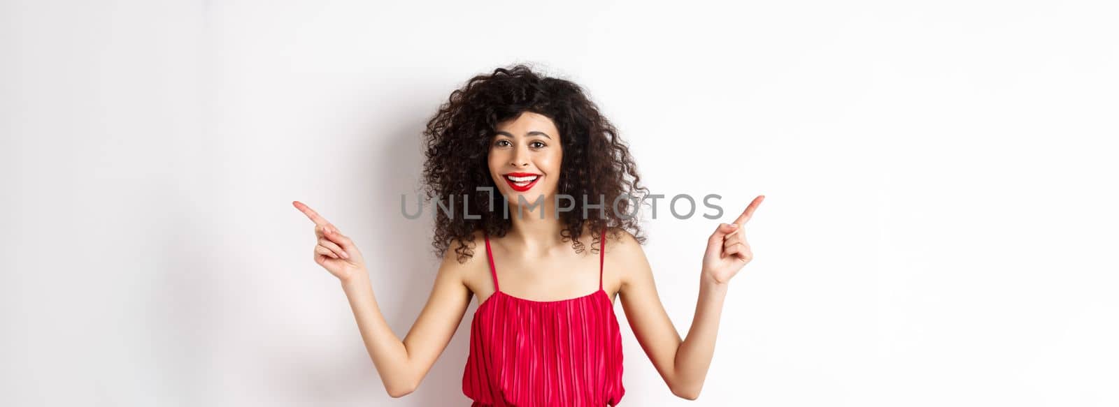 Happy lady with curly hairstyle and red lips, smiling white teeth, pointing sideways at two ways, showing advertisements aside, standing on white background.