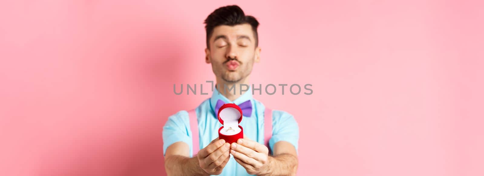 Valentines day. Silly boyfriend in bow-tie pucker lips, close eyes and waiting for kiss after making proposal, showing golden engagement ring in small red box, standing on pink background.