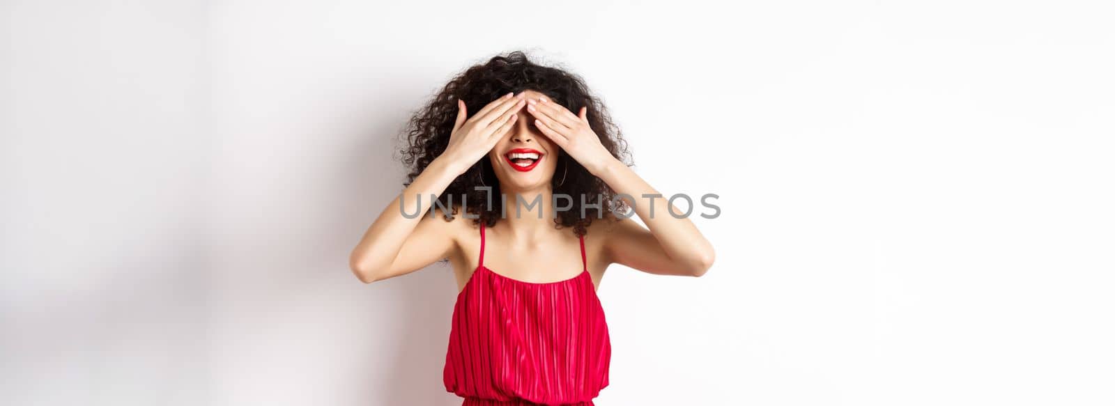 Excited curly-haired woman in party dress, close eyes and smiling with red lips, waiting for surprise with happy face, standing on white background.