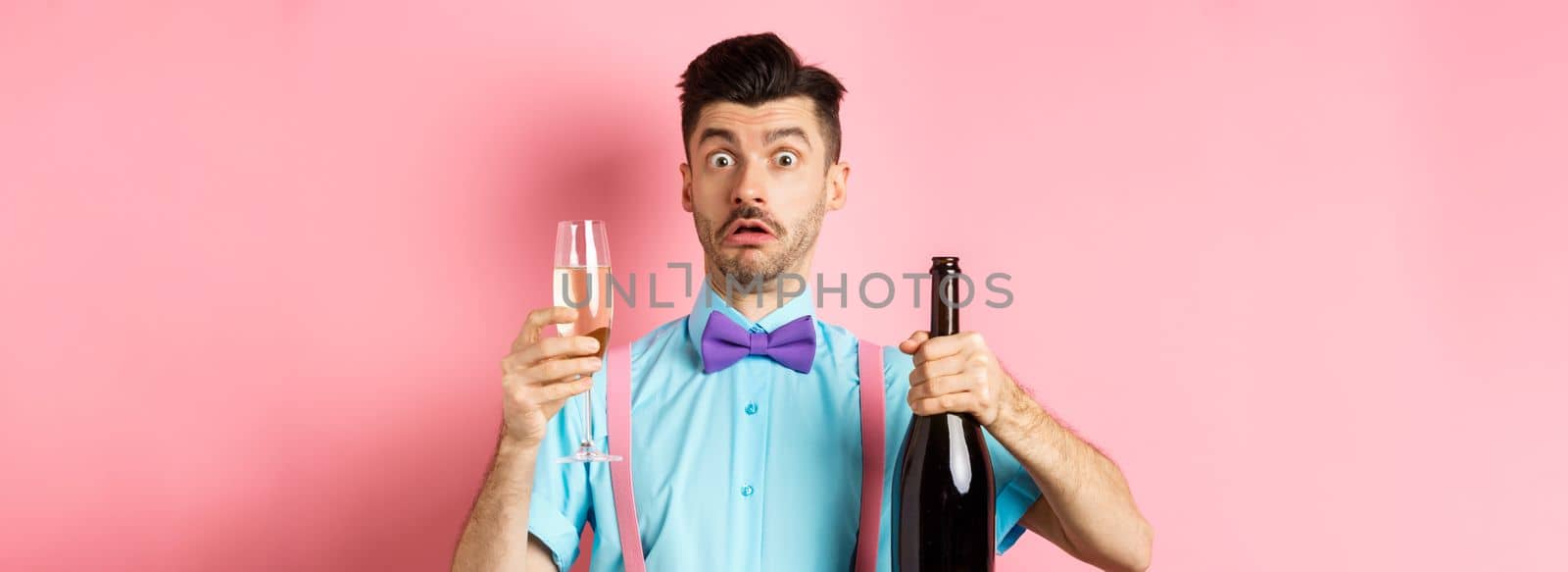 Holidays and celebration concept. Confused man staring startled at camera while pouring glass of champagne, standing puzzled over pink background.