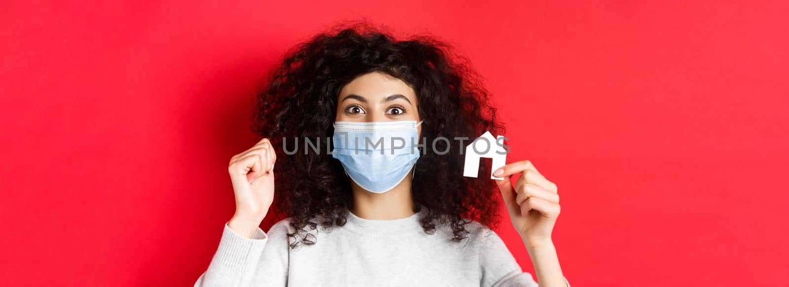 Real estate and covid-19 concept. Excited woman in medical mask showing small paper house cutout, standing on red background.