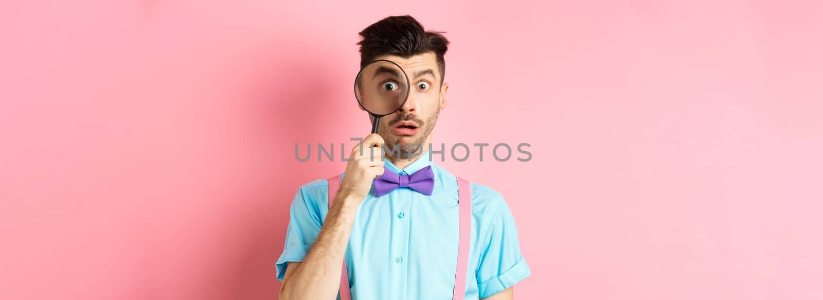 Funny guy look through magnifying glass with surprised face, seeing something interesting, standing on pink background.