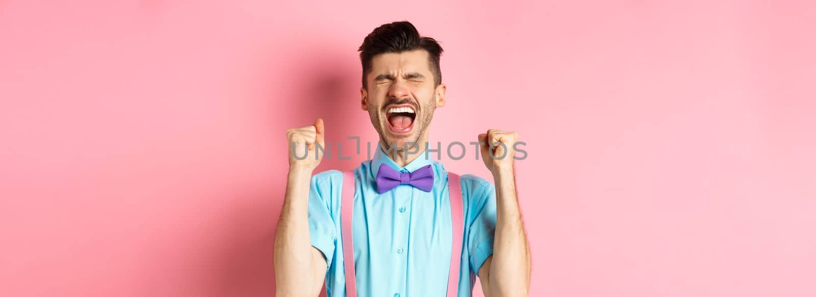 Relieved man shouting from happiness and joy, scream yes with closed eyes and clenched fists, celebrating victory, achieve goal and triumphing, standing over pink background.