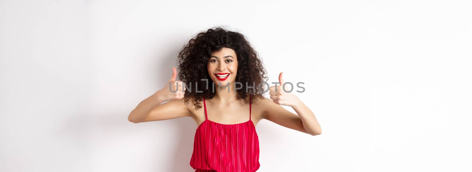 Attractive young woman recommending promo offer, showing thumb up and smiling, like product, standing in festive red dress on white background by Benzoix