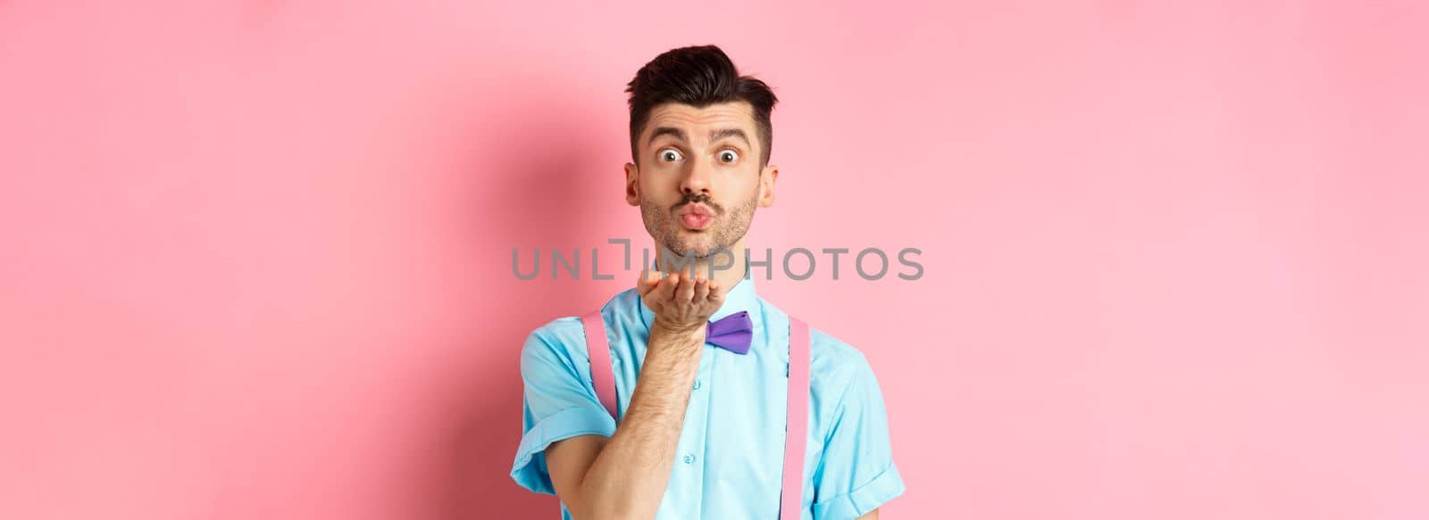 Cute boy sending air kiss at camera and saying I love you on Valentines day, standing over romantic pink background in bow-tie and shirt by Benzoix