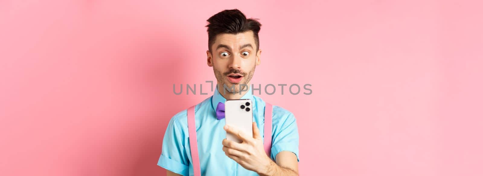 Impressed man reading mobile screen with amazed face, seeing online promo offer, standing over pink background. Copy space