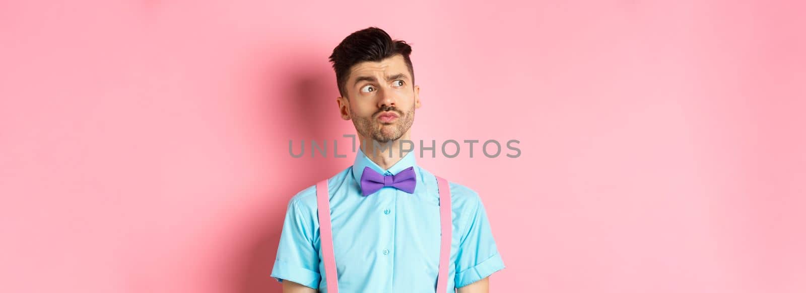 Pensive young man in romantic outfit, looking away and thinking, standing thoughtful on pink background in fancy bow-tie and shirt.
