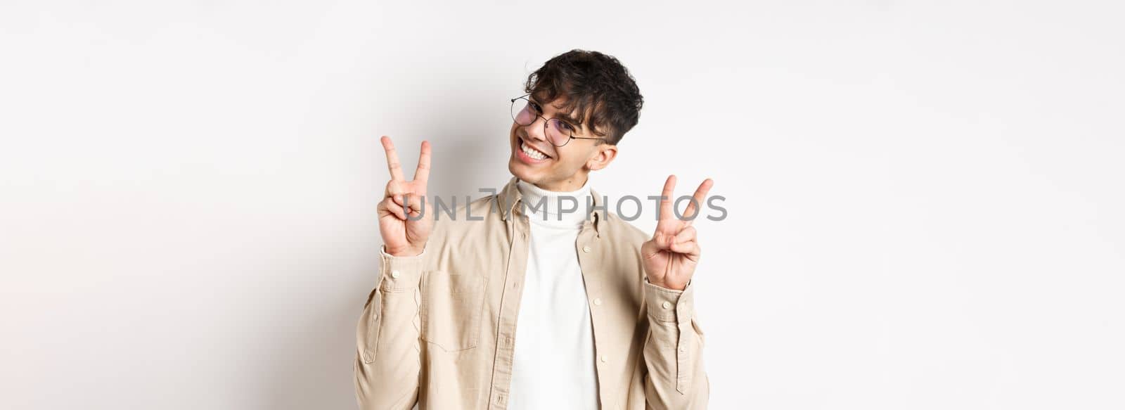 Real people. Handsome hipster guy in glasses showing peace gestures and smiling cute, standing on white background.