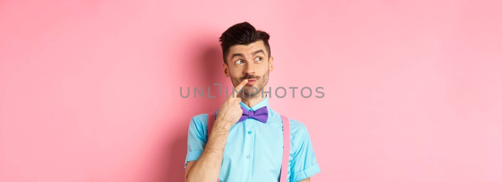 Image of handsome funny man in bow-tie looking thoughtful left and touching lip, thinking what to choose, making decision, standing over pink background.