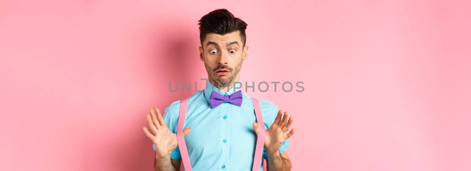 Funny guy with moustache and bow-tie, adjusting his suspenders and looking down with confused and surprised face, standing over pink background.