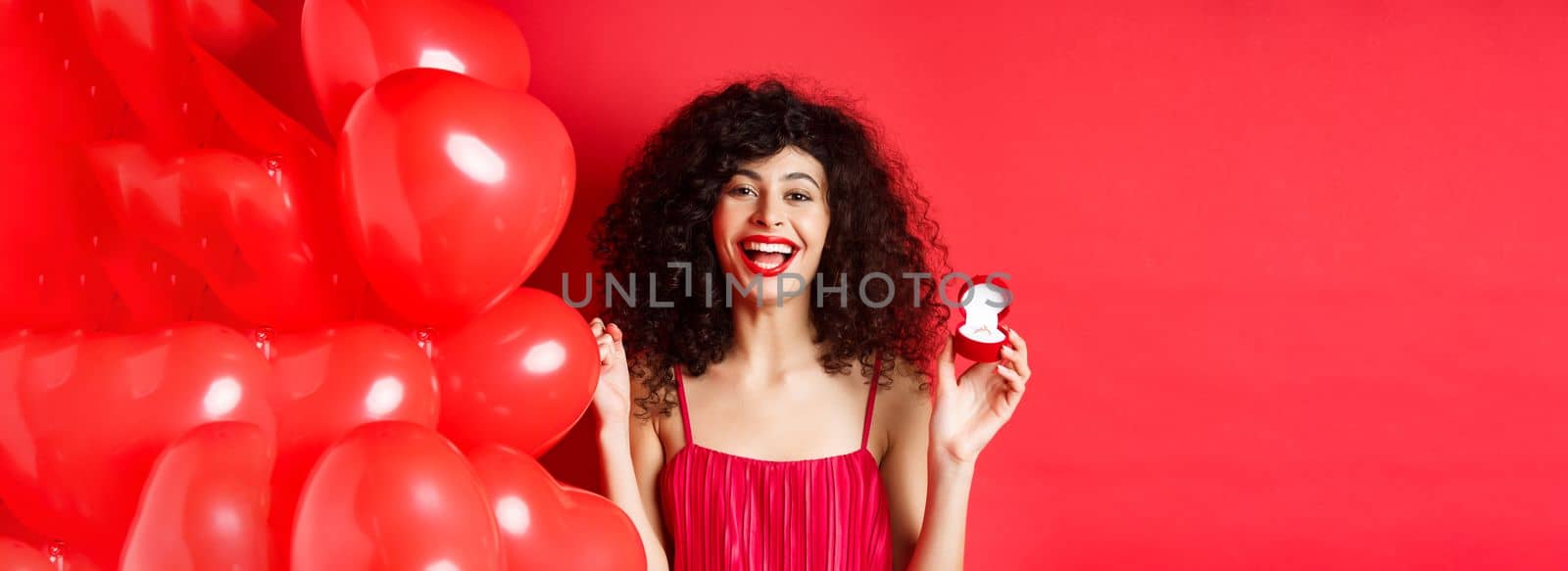 Happy beautiful lady feeling excited about marriage proposal, showing engagement ring and laughing, standing near heart balloons on red background by Benzoix