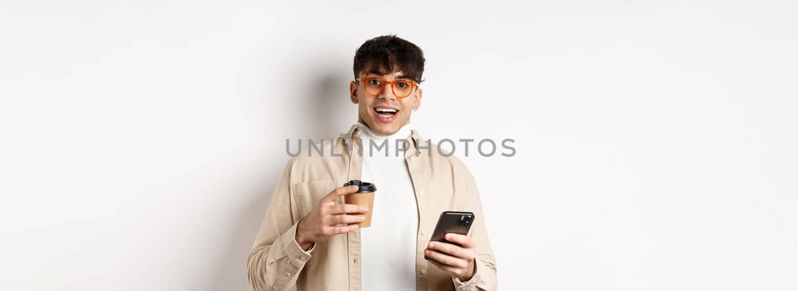 Cheerful smiling young man in glasses drinking coffee and holding smartphone, looking excited at camera, standing on white background by Benzoix