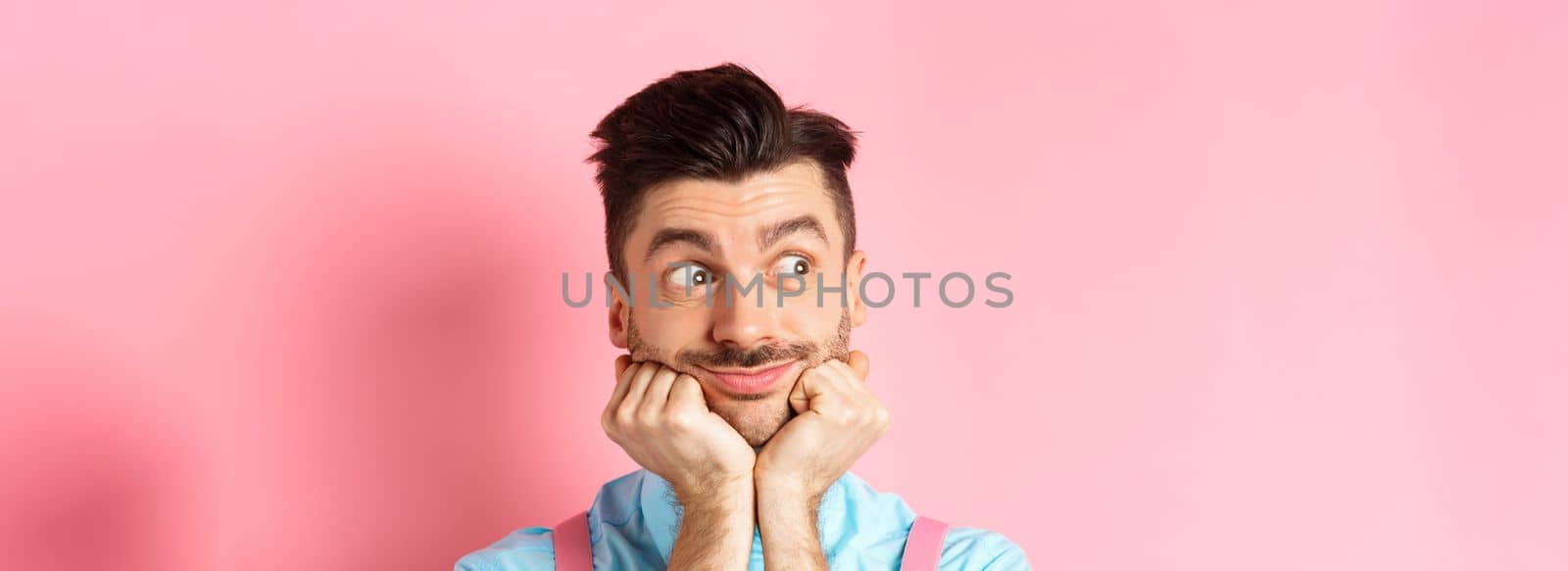 Close up of cute guy dreaming of something, looking aside and imaging things, standing on pink background, leaning face on hands, pink background.