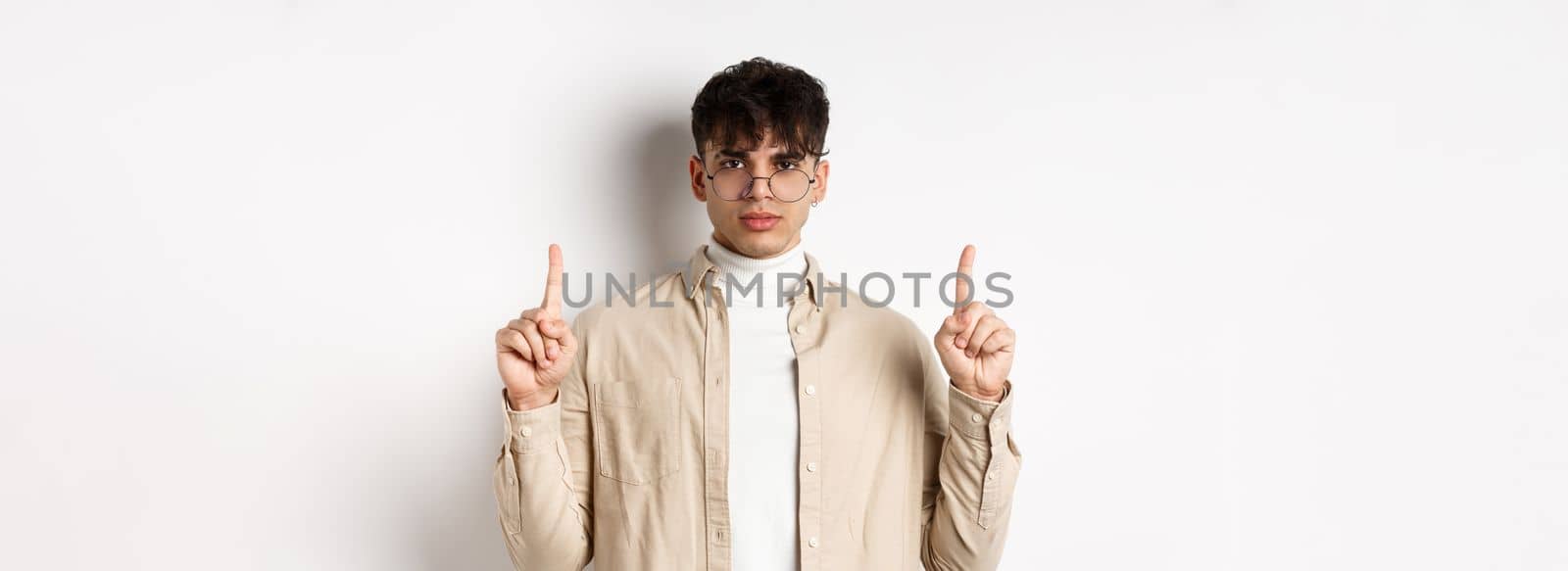 Handsome hipster guy in glasses pointing fingers up at empty space, looking serious at camera, standing on white background.