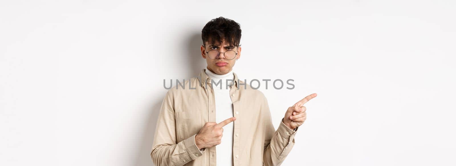 Sad and grumpy boy pointing fingers left, sulking with childish expression, complaining on something bad, standing on white background.