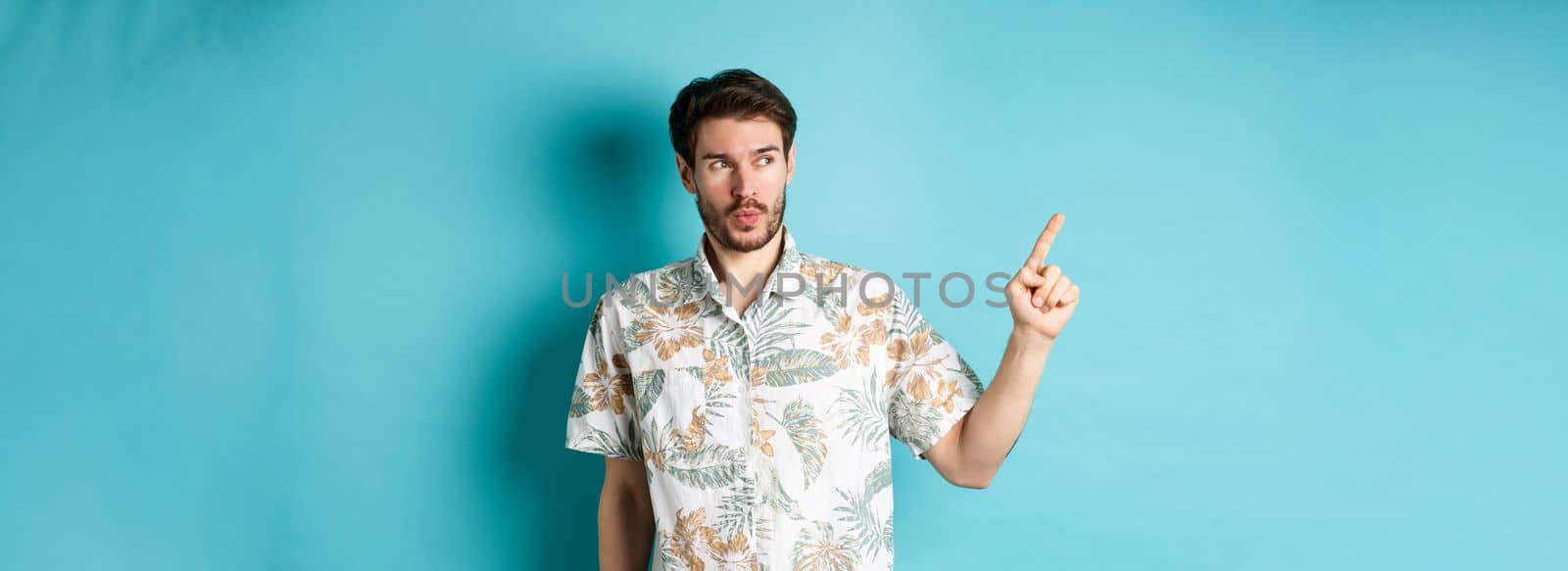 Handsome guy wearing summer shirt on vacation, pointing and looking aside at empty space, making amazed face, standing on blue background.