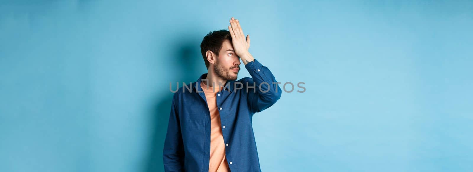 Annoyed and tired man facepalm, roll eyes and look bothered by stupid behaviour, standing on blue background.