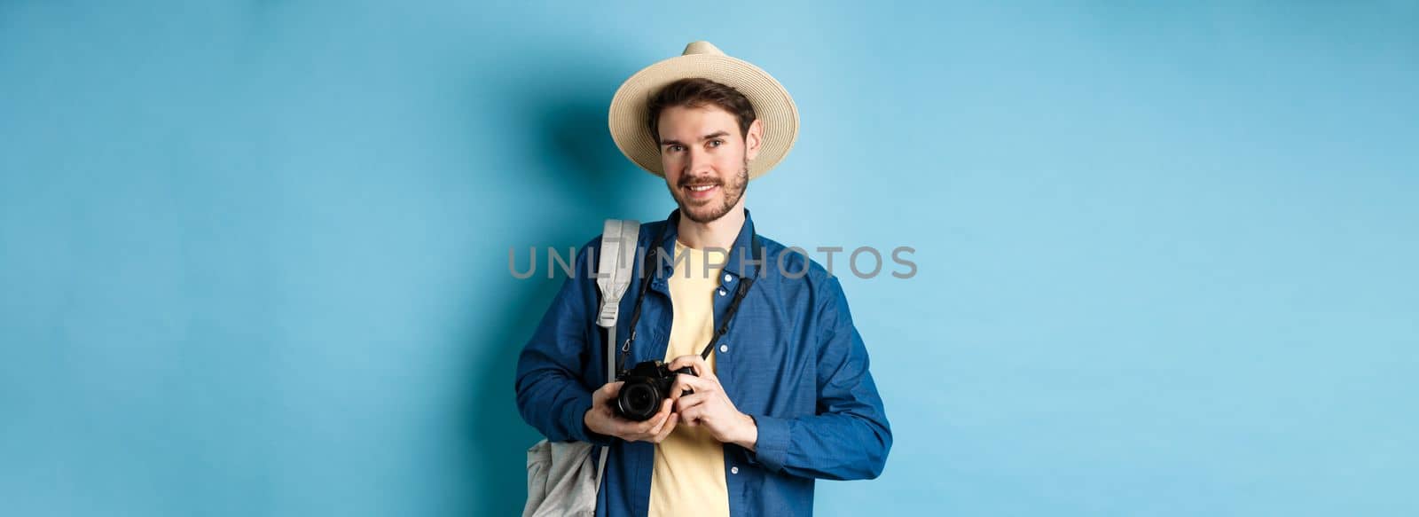 Handsome guy going on summer vacation, backpacking on holiday. Tourist with straw hat and camera smiling happy, standing on blue background.