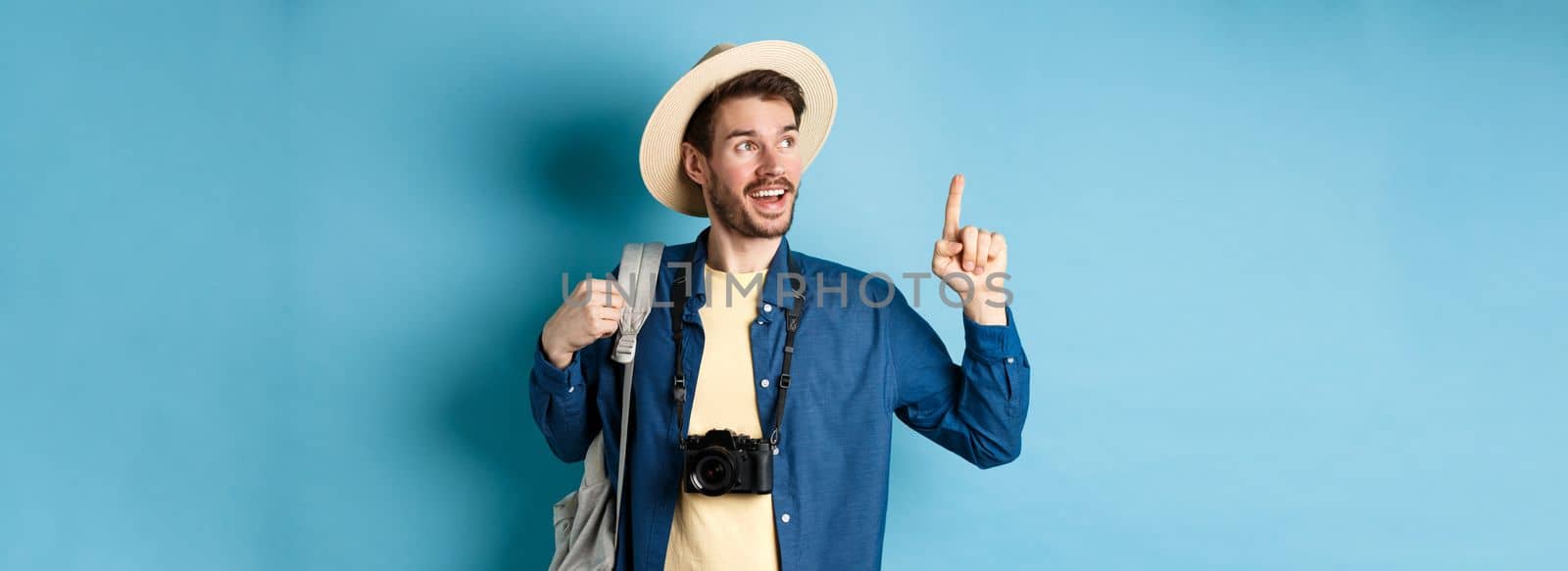 Handsome tourist looking and pointing up, travelling on summer vacation with backpack and camera, standing on blue background.