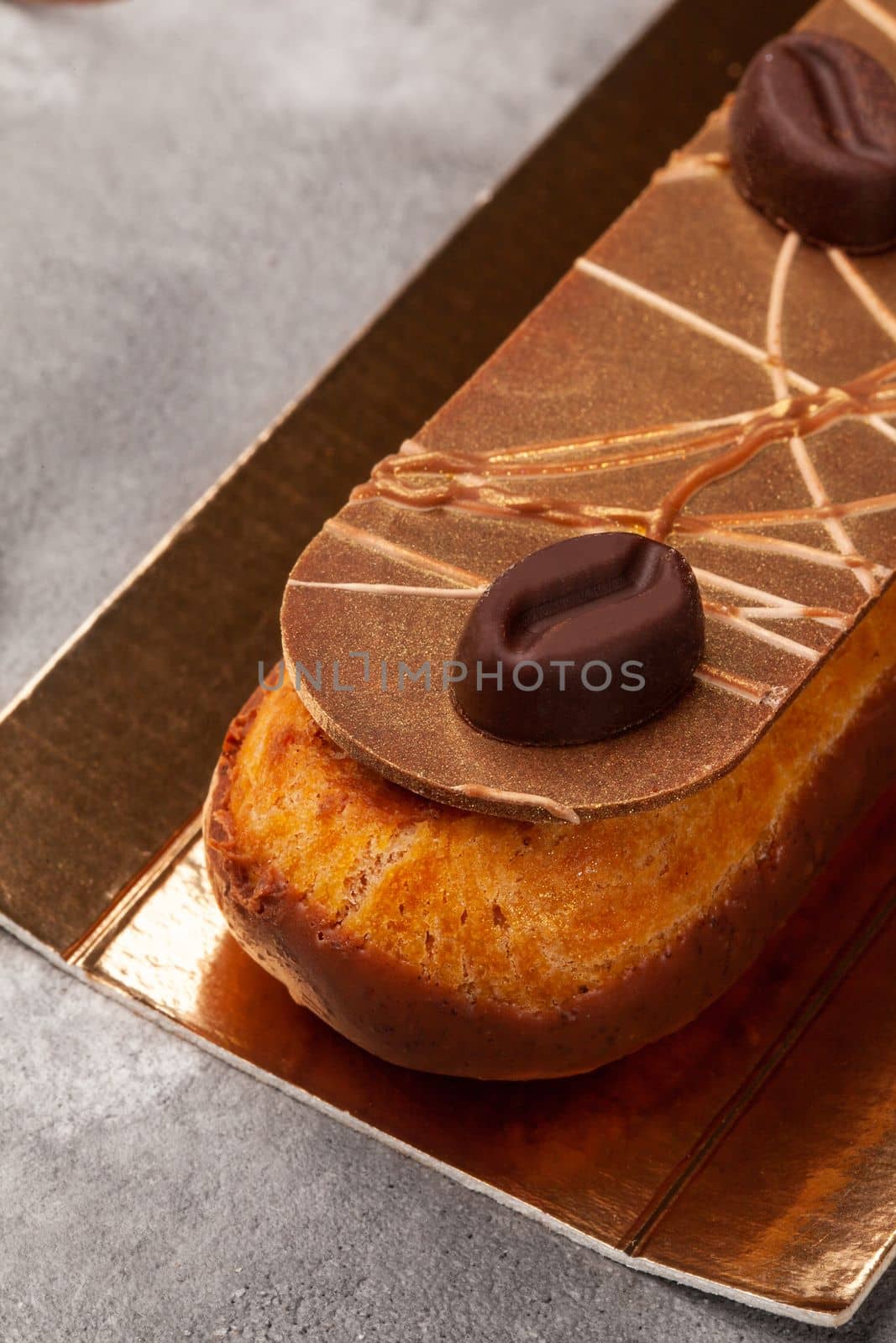 Closeup of appetizing eclair with custard filling, chocolate icing and chocolate coffee beans on gold cardboard on gray background, cropped shot. Traditional French choux dough pastry