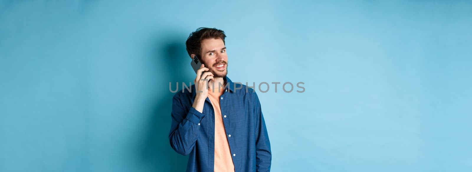 Modern smiling guy talking on mobile phone, looking happy at camera, standing on blue background by Benzoix