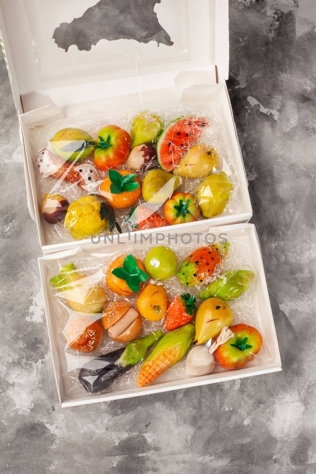 Various of colorful marzipan sweets in form of fruits and vegetables in open carton boxes on gray surface. Popular Italian treats traditionally made of almond paste. Craftsmanship in confectionery art